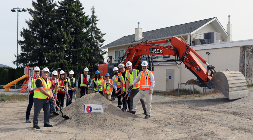 Mayor and Council and others pose with shovels in their hands behind a mound of dirt, in a groundbreaking photo in front of an excavator and the old Lakeview Community Centre